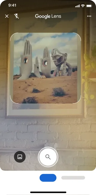 Auf einem Handy ist die Lens App geöffnet. Um nach einem Bild von großen steinernen Handskulpturen in einer Wüste zu suchen, kann auf den Auslöser getippt werden.
